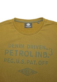 Olíva zöld pamut póló, kerek nyakkivágással, rövid ujjakkal és elülső nyomattal: "DENIM DRIVEN PETROLIND.3 REG. U.S. PAT. OFF."