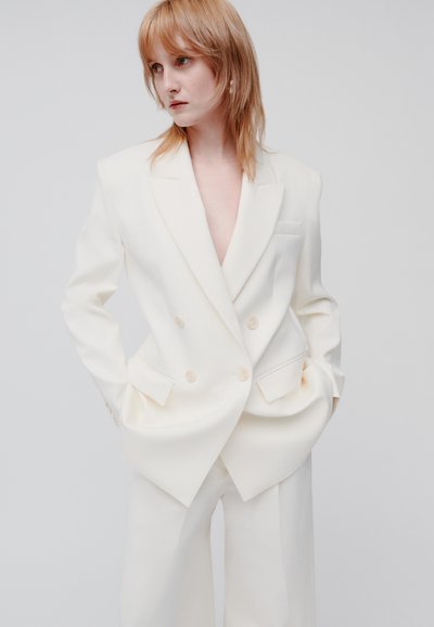 Witte dubbelgebreide blazer met een diepe V-hals, oversized pasvorm, geknipte revers, zijzakken en lichte knoopaccenten, gecombineerd met bijpassende trousers.
