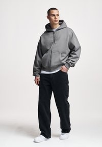 Joven de pie con una sudadera gris con cremallera, vaqueros negros, zapatillas blancas, con la mano izquierda en el bolsillo, mirando hacia un lado.