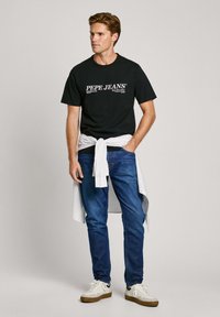 Melnas kokvilnas T-krekls ar "PEPE JEANS" logo, zili džinsu bikses un baltas kedas. Ap vidukli sasiets svītrains krekls piešķir papildus detaļas.