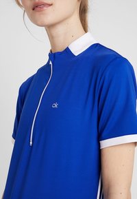 Niebieska koszulka polo wykonana z gładkiego materiału, z białym kołnierzykiem i zamkiem błyskawicznym, z małym białym logo "ck" na przedzie. Krótkie rękawy.