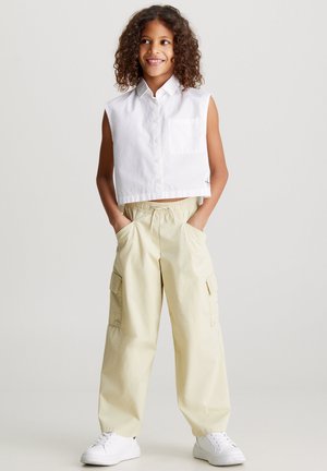 Bambino con capelli ricci che indossa una camicia bianca senza maniche, pantaloni cargo beige con le mani nelle tasche e sneakers bianche, in piedi e sorridente.