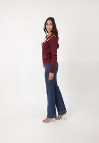 Suéter burdeos con detalle de cuello blanco, combinado con jeans azules acampanados y sandalias de tacón transparente. Textura suave y diseño ajustado.