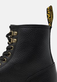 Černá kožená kotníčková bota s texturovaným povrchem, s zlatými průvleky, černými tkaničkami a žlutým poutkem na zadní straně.