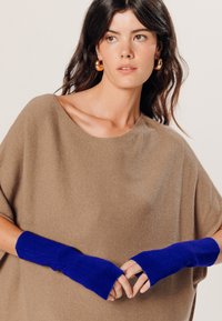 Bruine, ribbelige gebreide poncho met een brede halslijn, gecombineerd met lange, levendig blauwe vingerloze handschoenen. De textuur lijkt zacht en gezellig.
