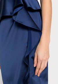 Mörkblå klänning med en slät satintextur, lager av volanger på bodicen och en figurnära silhuett.