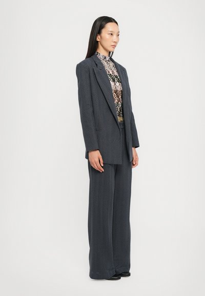 Completo in grigio scuro a spina di pesce su misura con blazer oversize e pantaloni a gamba larga, indossato sopra un top a collo alto floreale. Tessuto morbido, design strutturato.