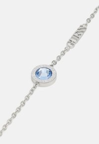 Bracciale in argento con un pendente circolare e una pietra azzurra chiara, inciso con la parola "GUESS," attaccato a una chiusura elegante.
