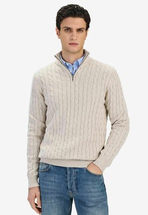 Mand iført beige kabelstrikket halv-zip sweater over blå skjorte med krave og blå jeans, den ene hånd i lommen, står mod hvid baggrund.