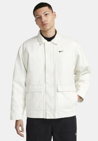 Chaqueta blanca de Nike con diseño tipo collar, bolsillos frontales y un logo sutil en el lado izquierdo del pecho. El material parece texturizado y resistente.