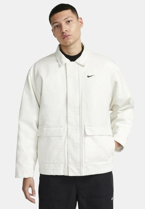 White Nike jacket with a collared design, front pockets, and a subtle logo on the left chest. Material appears textured and sturdy.