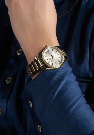 Orologio d'oro con quadrante argentato, bracciale testurizzato e lunetta adornata da cristalli. Indossato su un polso, abbinato a una camicia blu testurizzata.