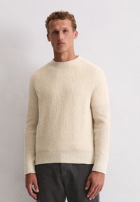 Beige gestrickter Pullover mit Rippstruktur, Rundhals und langen Ärmeln. Kombiniert mit dunklen, leicht strukturierten Hosen.