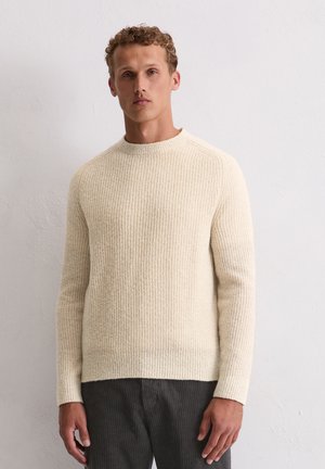 Beige gebreide trui met een geribbelde textuur, ronde hals en lange mouwen. Gepaard met donkere, licht getextureerde broek.