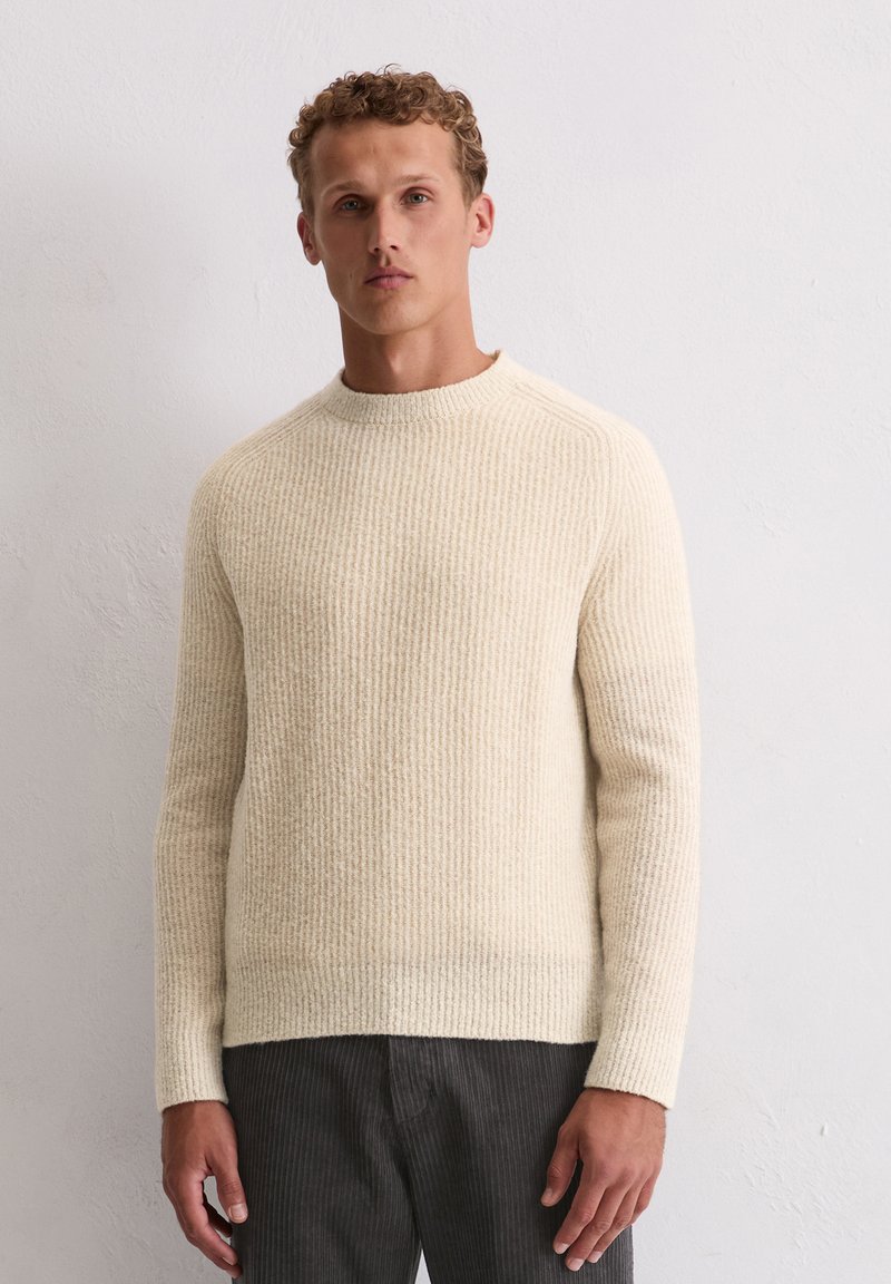 Beige gestrickter Pullover mit Rippstruktur, Rundhals und langen Ärmeln. Kombiniert mit dunklen, leicht strukturierten Hosen.