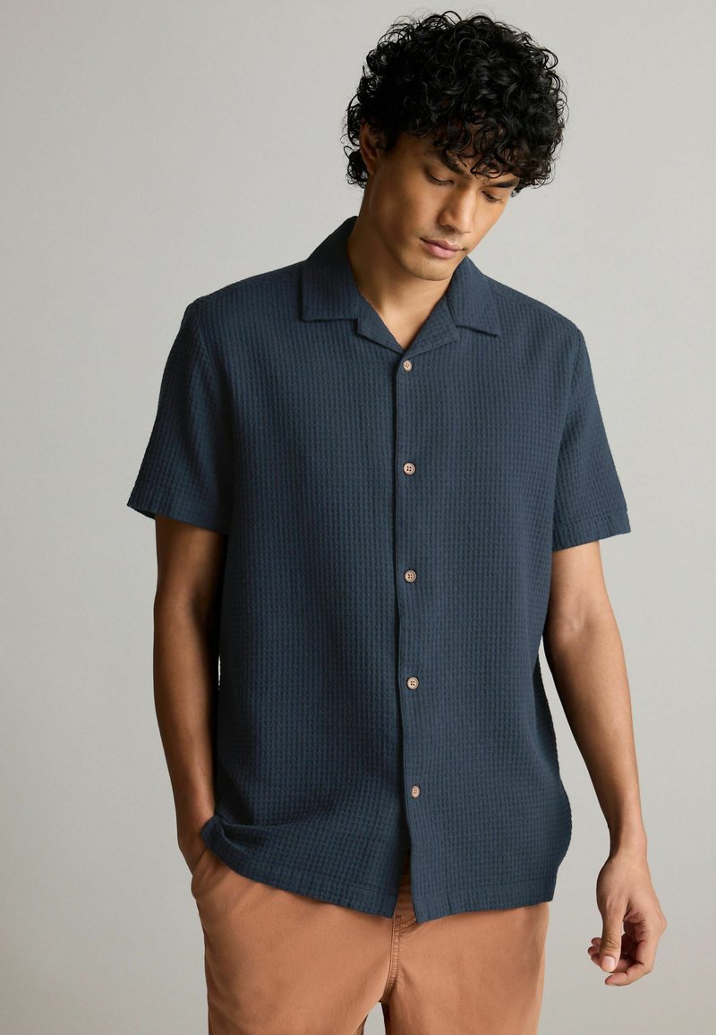 Camisa de manga corta, con botones, en tela texturizada azul oscura, con cuello, presenta botones marrones y un corte holgado.