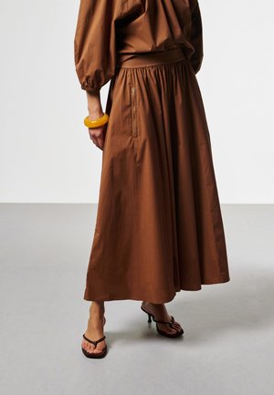 Vrouw draagt een bruine enkellange rok met ritszak, bijpassende blouse, geel armbandje en zwarte sandalen met hak en bandjes.