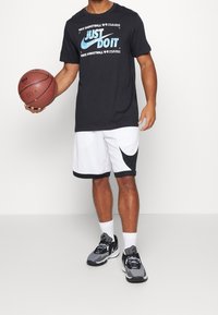 Camiseta negra de manga corta con estampado azul de "Just Do It", pantalones cortos de baloncesto blancos con detalles negros y zapatillas deportivas grises; sosteniendo un balón de baloncesto.