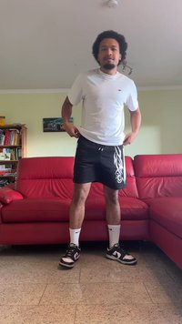 Weiße Kurzarm-T-Shirt, schwarze Shorts mit weißem tribal Design, weiße Socken, schwarz-weiße Turnschuhe, stehend in einem Wohnzimmer.