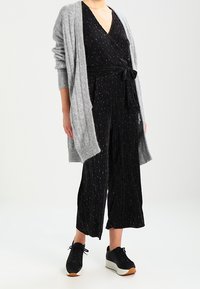 Svart wrap jumpsuit med silverränder, knuten i midjan; kombinerad med en grå stickad cardigan och svarta sneakers med vita sulor.