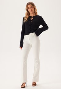 Haut court en tricot côtelé noir avec détail à nouer, associé à un pantalon blanc évasé. Le modèle porte des talons noirs à lanières, mettant en valeur une silhouette élégante.