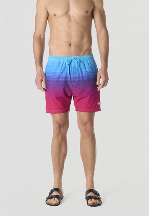 Hombre sin camisa que lleva un bañador de gradiente de azul a rojo con cordón y sandalias negras sin talón, de pie contra un fondo liso.