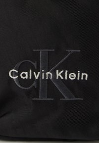 Fekete vászontáska, amelyen világosszürke hímzett "Calvin Klein" logó látható, a "CK" kezdőbetűkkel kiemelten megjelenítve.