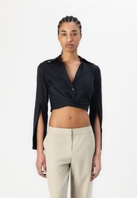 Crna skraćena majica s dugim rukavima, s bočnim prorezima i zlatnim akcentima na ramenima, u kombinaciji s svijetlosmeškim hlačama.