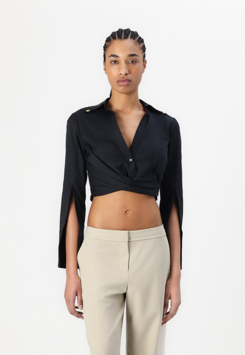 Crna skraćena majica s dugim rukavima, s bočnim prorezima i zlatnim akcentima na ramenima, u kombinaciji s svijetlosmeškim hlačama.