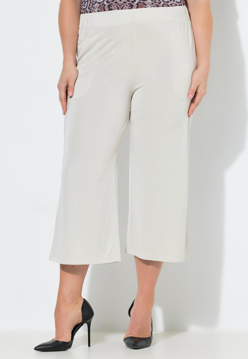 Femme portant un pantalon large écru cropped associé à des chaussures slingback noires à talons hauts, debout sur un fond blanc.