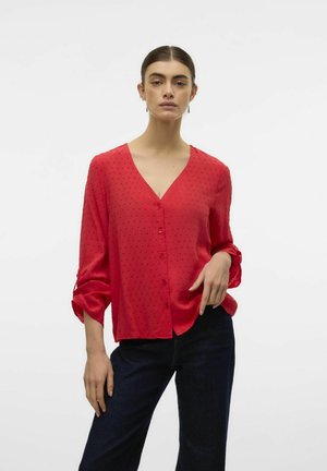 Blusa rossa con scollo a V, frontale con bottoni e tessuto testurizzato caratterizzato da delicati punti in rilievo. Abbinata a jeans blu scuro.