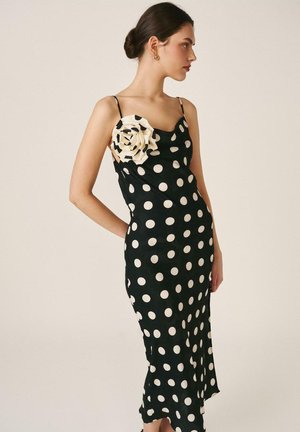 Mujer con un vestido negro sin mangas con lunares blancos y una gran flor de tela color crema en el pecho, de pie de perfil.