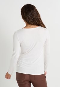 Haut à manches longues, blanc, en tissu doux et ajusté. Il présente un col rond et des coutures lisses et minimales. Porté avec un pantalon marron.