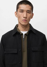 Black button-up jacket made of textured fabric, featuring two chest pockets and a collar, layered over a zippered olive green top.