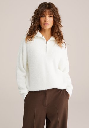 Mujer con cabello rizado lleva un suéter blanco de felpa con cremallera y pantalones marrones, de pie con las manos en los bolsillos contra un fondo neutral.