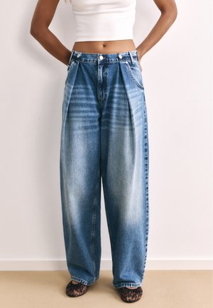 Kvinde iført højtaljede, vidde, blå jeans med falmet look, en hvid cropped tank top og leopardmønstrede sko, stående med hænderne på hofterne.