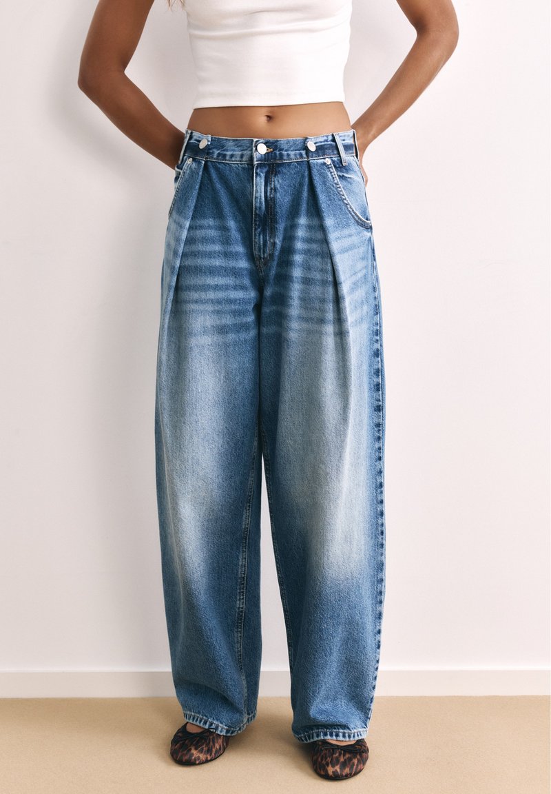 Femme portant un jean bleu délavé taille haute à jambes larges, un débardeur court blanc et des chaussures à motif léopard, debout les mains sur les hanches.