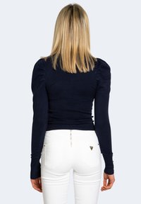 Femme blonde portant un haut ajusté bleu marine à manches longues avec des épaules bouffantes et un pantalon blanc, de dos contre un fond uni.