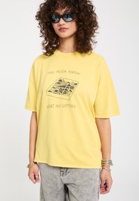 Femme aux cheveux bouclés portant des lunettes de soleil noires, un t-shirt jaune avec des chocolats et le texte « On ne sait jamais ce qu'on va obtenir », et un jean gris clair.