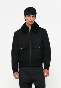 Chaqueta bomber negra con un suave cuello de piel negro, cierre de cremallera en el frente, dos bolsillos frontales con cierres a presión y puños y dobladillo acanalados.