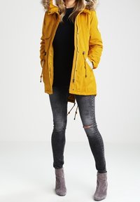 Žlutá parka s kapucí podšitou kožešinou, zapínání na zip a druky, stažený pas, nošena přes černý top a černé obnošené džíny, doplněna šedými kotníkovými botami.