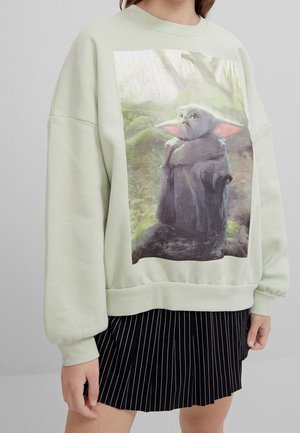 Persoon draagt een lichtgroene sweater met een bedrukte afbeelding van een klein groen buitenaards figuurtje in een bosomgeving, gecombineerd met een zwarte geplooide pinstripe rok.