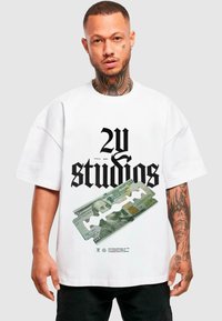 Balta pārāk liela T-kreklu ar drosmīgu melnu "2U studios" tekstu un drukātu simts dolāru rēķina grafiku. Īsas piedurknes, brīva piegriezuma.