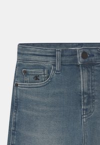 Denim jeans in medium blue, featuring a front pocket with a black logo, a button closure, and contrast stitching along the seams.