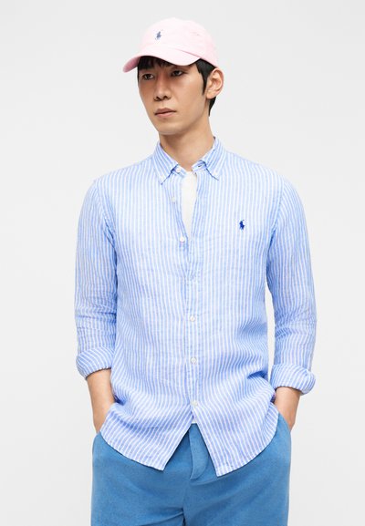 Άνδρας που φορά ροζ καπέλο, πουκάμισο με κάθετες ρίγες σε γαλάζιο και άσπρο, λευκό εσωτερικό μπλουζάκι και μπλε παντελόνι, στέκεται με τα χέρια στις τσέπες.