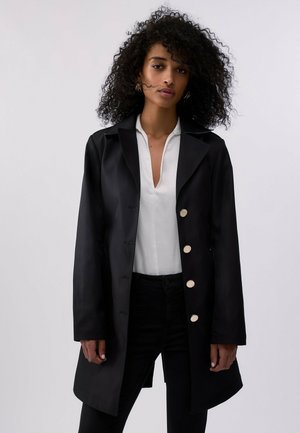 Donna con capelli ricci che indossa un cappotto nero con bottoni dorati sopra una camicetta bianca con scollo a V e pantaloni neri su uno sfondo chiaro e uniforme.