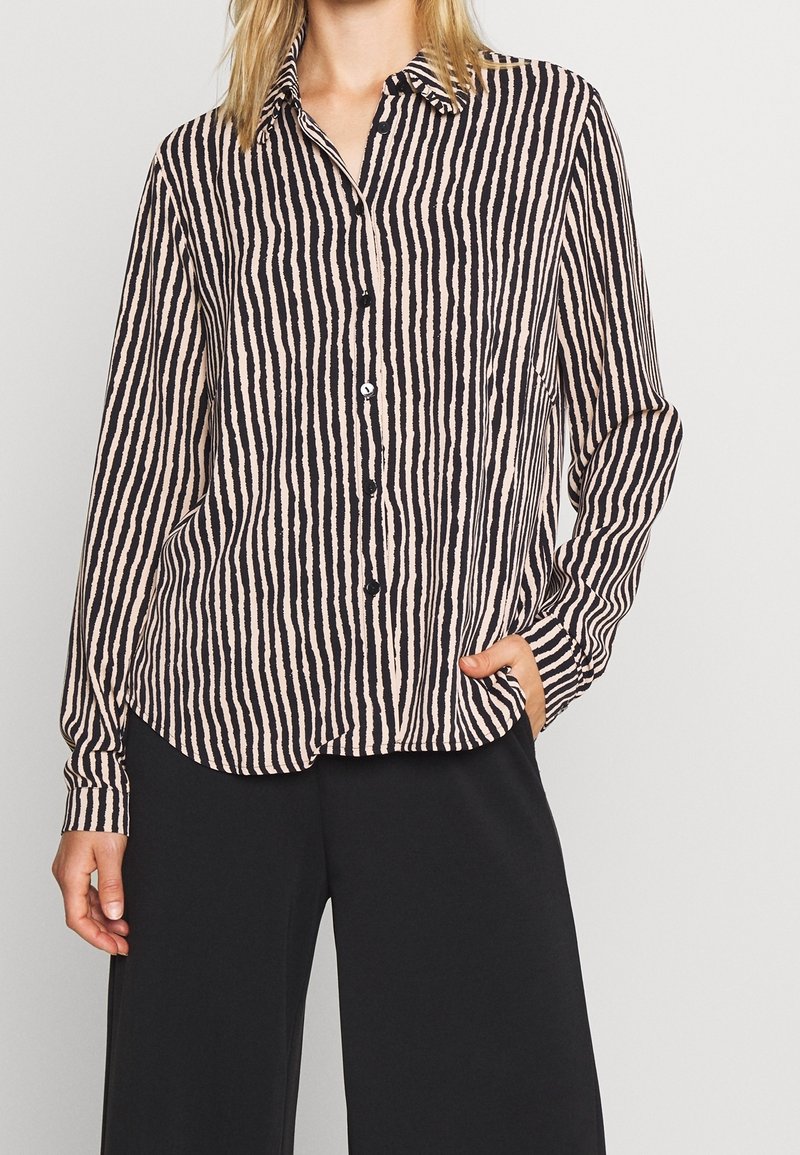 Femme portant une chemise à manches longues rayée verticalement noir et beige, boutonnée, avec un pantalon large noir, une main dans la poche.