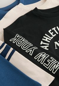 Trys apykaklės megztiniai, sukrauti vienas ant kito: mėlynas tekstūruotas audinys, bežinis lygaus audinio ir juodas su balta spauda "NEW YORK" bei dryžuotais akcentais.
