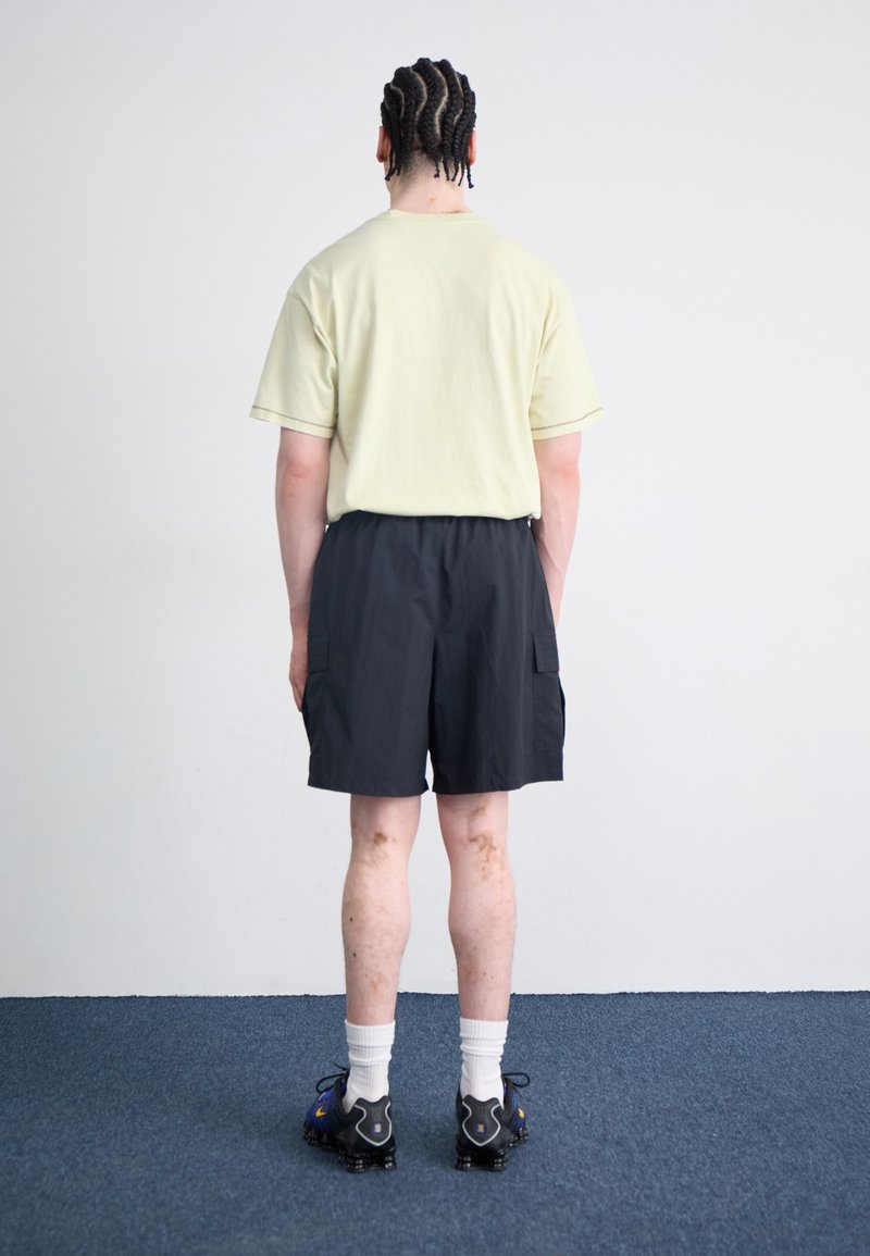Hellgelbes T-Shirt, schwarze Cargo-Shorts mit Seitentaschen, weiße Socken, Crew-Länge und schwarze Sportschuhe mit blauen Akzenten. Einfarbiger Hintergrund.