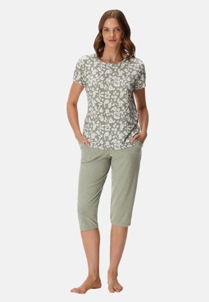 Mujer de pie descalza con una blusa de manga corta con estampado floral y pantalones cortos de color verde claro con las manos en los bolsillos sobre fondo blanco.